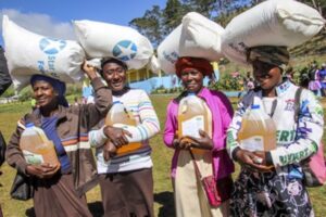 A group of four adult women holding jugs of cooking oil and balancing a sack of rice of their heads.